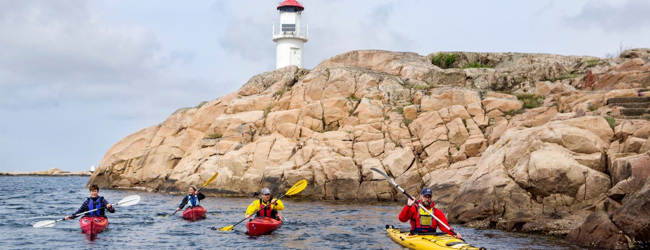 Ett gäng personer som paddlar kajak med en fyr i bakgrunden.