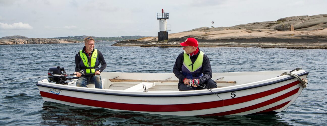 Två personer i en fiskebåt med en fyr i bakgrunden.