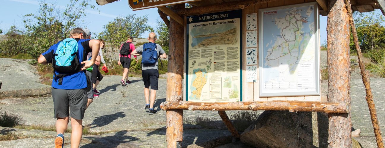 Ett gäng vandrare som går förbi en stor informations-skylt på Ramsvik i Bohuslän på västkusten.