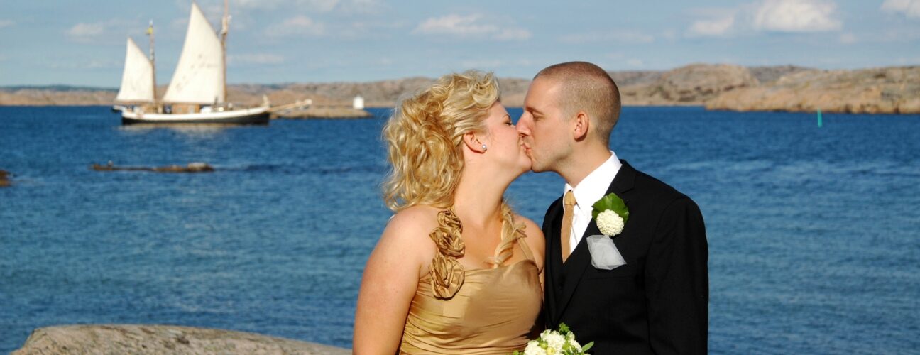 En kvinna och en man i bröllopskläder som kysser varandra med havet i bakgrunden.